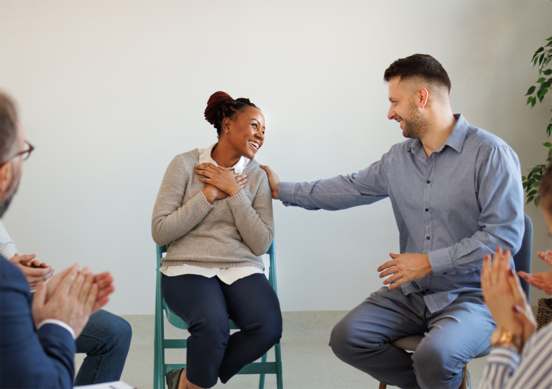 woman smiling while man putting hand on her shoulder and people around clapping